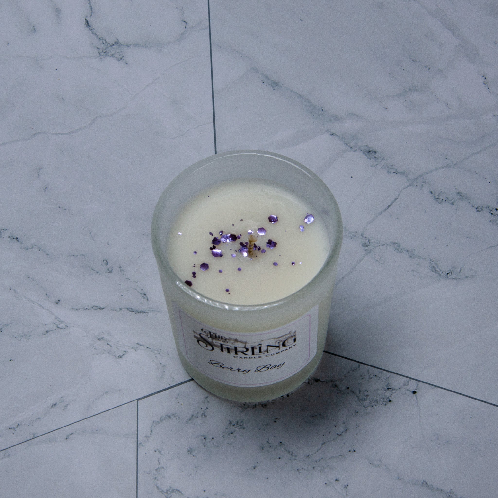 A clear glass candle jar with white wax and purple glitter labeled stirling candle company berry bay small candle