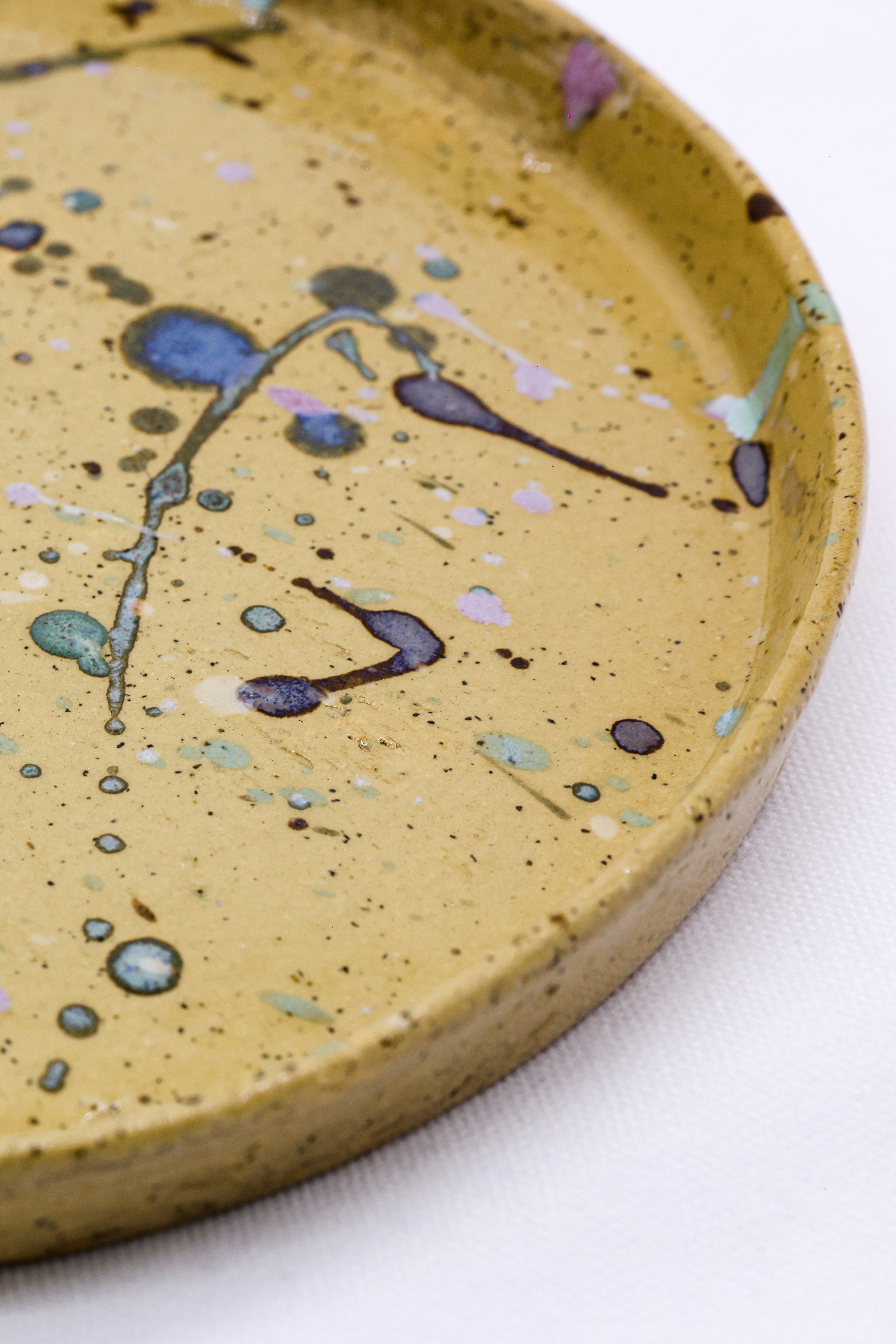 A yellow ceramic dinner plate with speckled texture and colorful paint splatters in blue purple green and pink