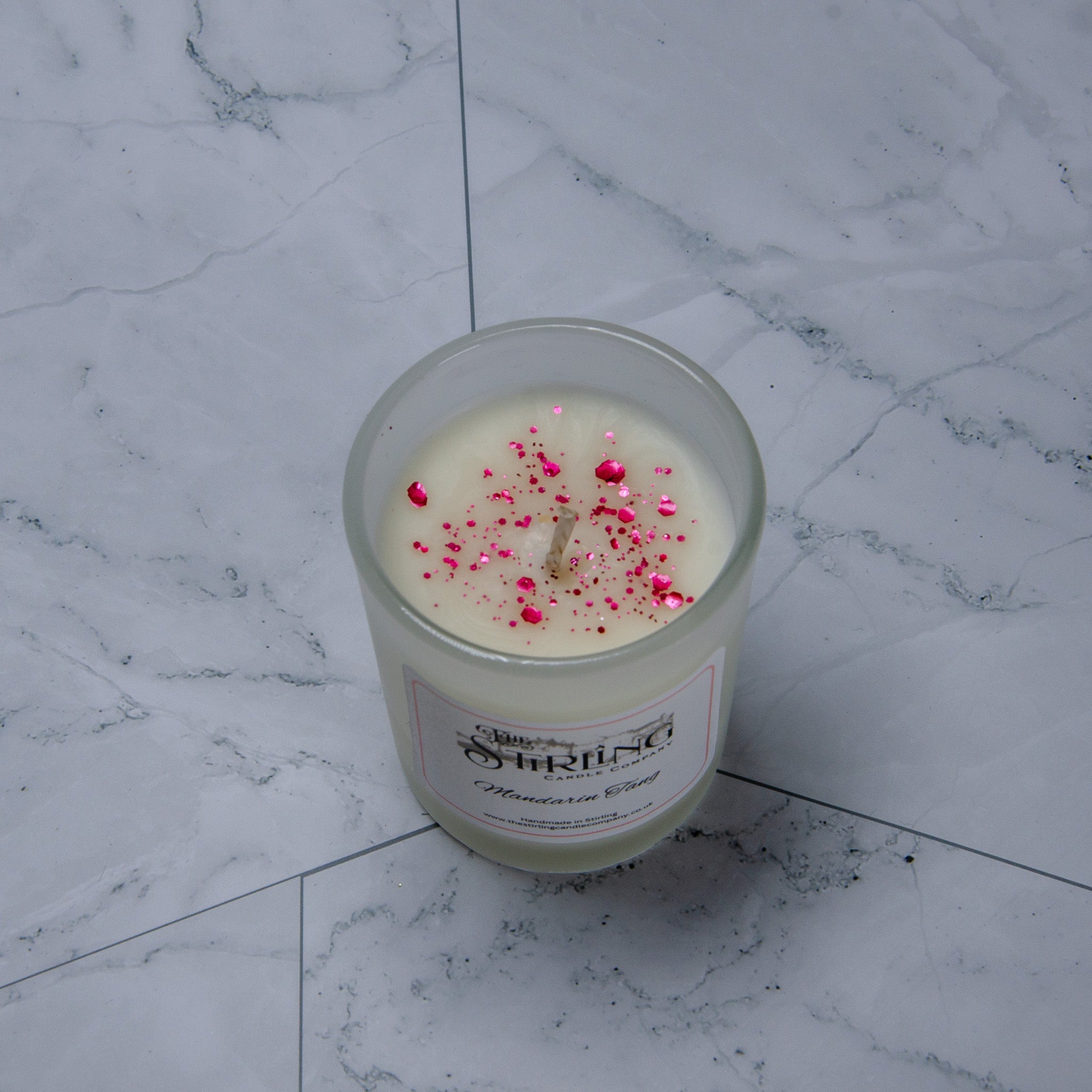 A creamy white mandarin tang scented candle in a clear glass jar topped with pink glitter by the shiring candle co