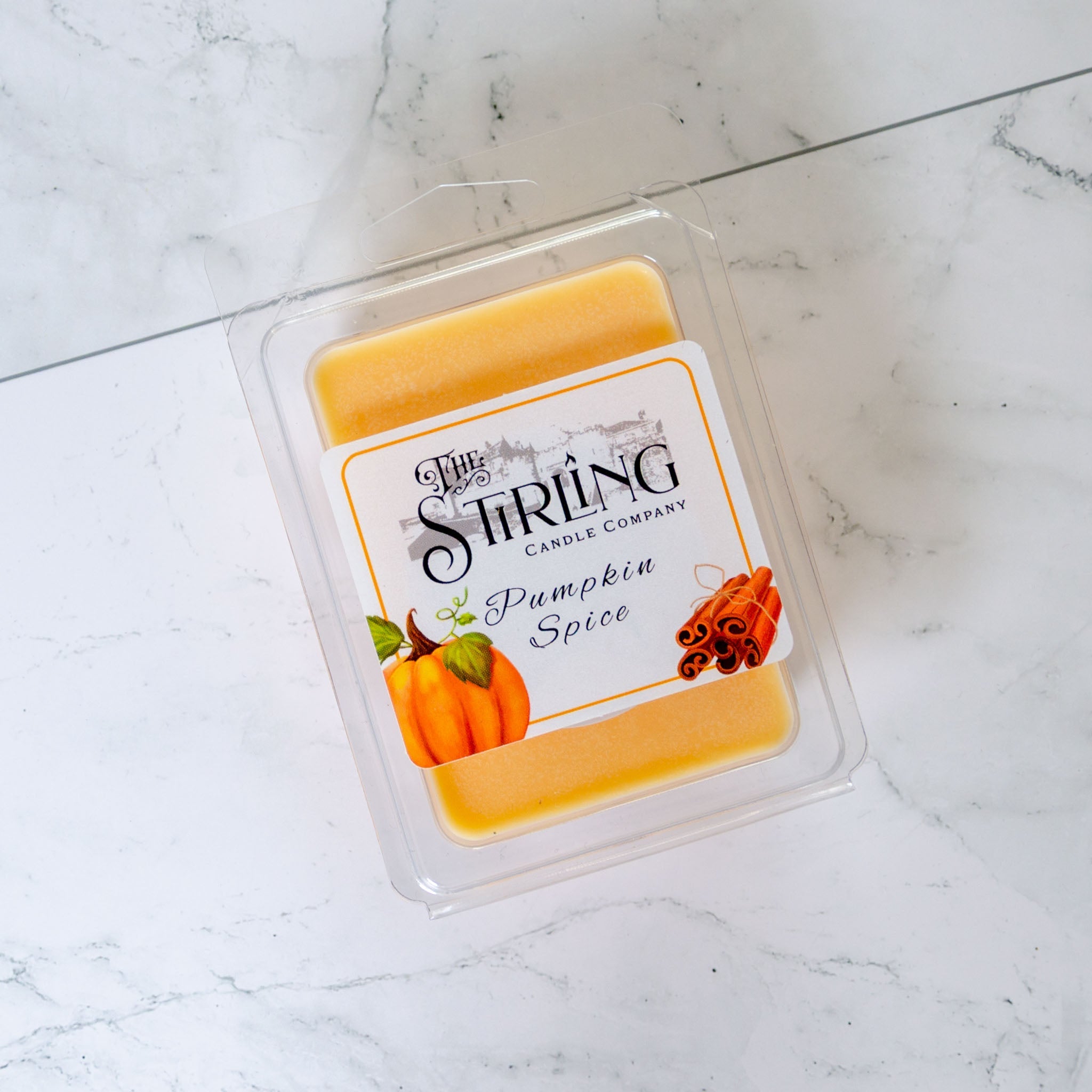 A golden-orange pumpkin spice wax melt clamshell in a clear plastic container with a white label showing pumpkins and cinnamon sticks