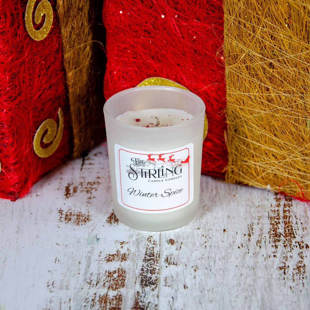 White candle in a frosted glass container labeled "Winter Spice" surrounded by red and gold decorative items.