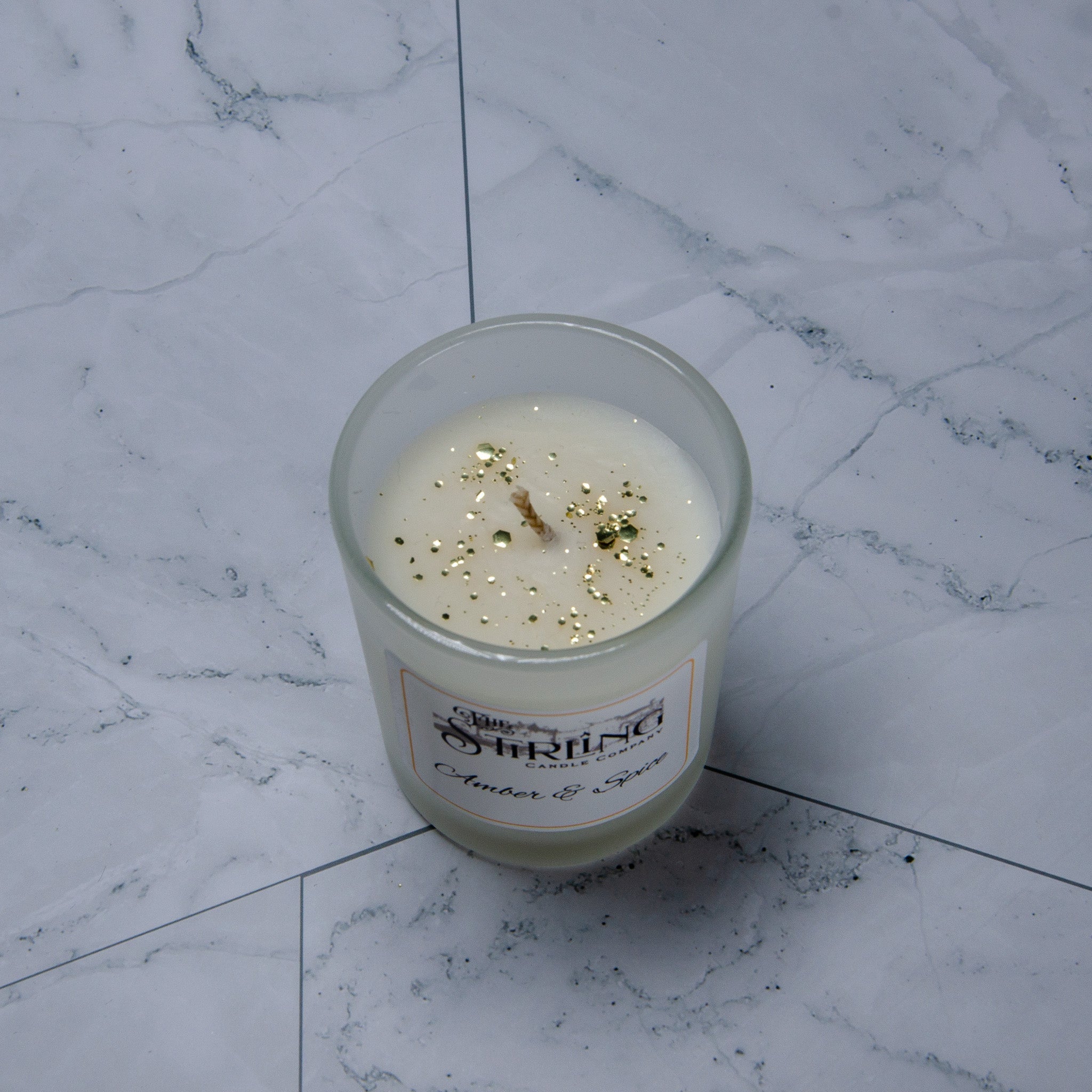 A creamy white scented candle in a clear glass jar with golden glitter labeled stirling candle co amber and spice small candle