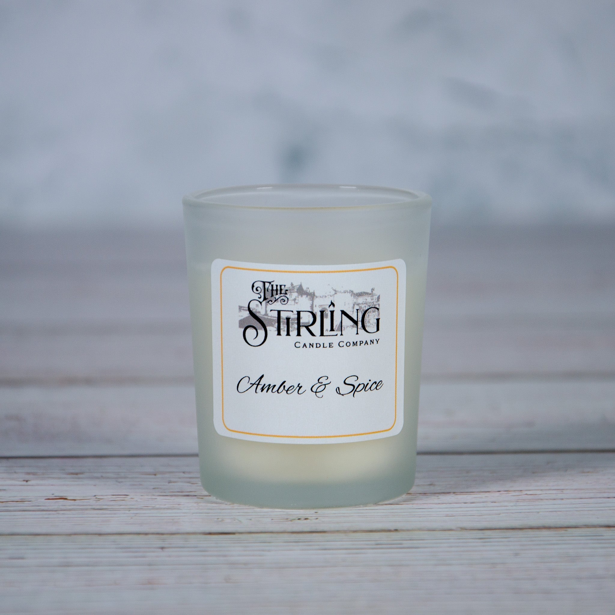 A frosted glass candle jar with a white label featuring black text and illustration amber and spice scent by stirling candle company