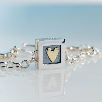 Silver square pendant bracelet with a gold heart inset on a textured gray background heart of gold bracelet