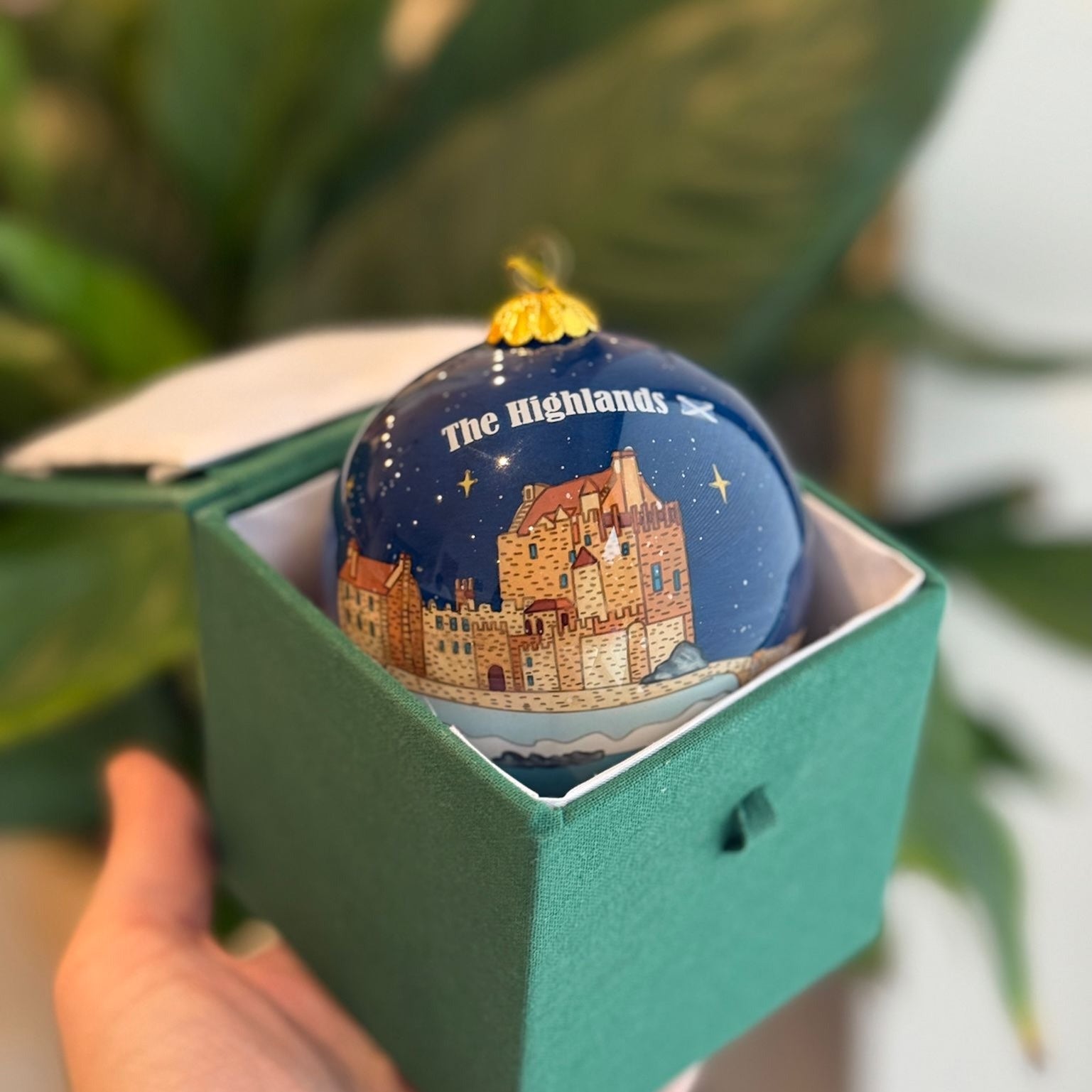 A glossy navy blue glass christmas bauble with a castle illustration and the text the highlands topped with a gold cap