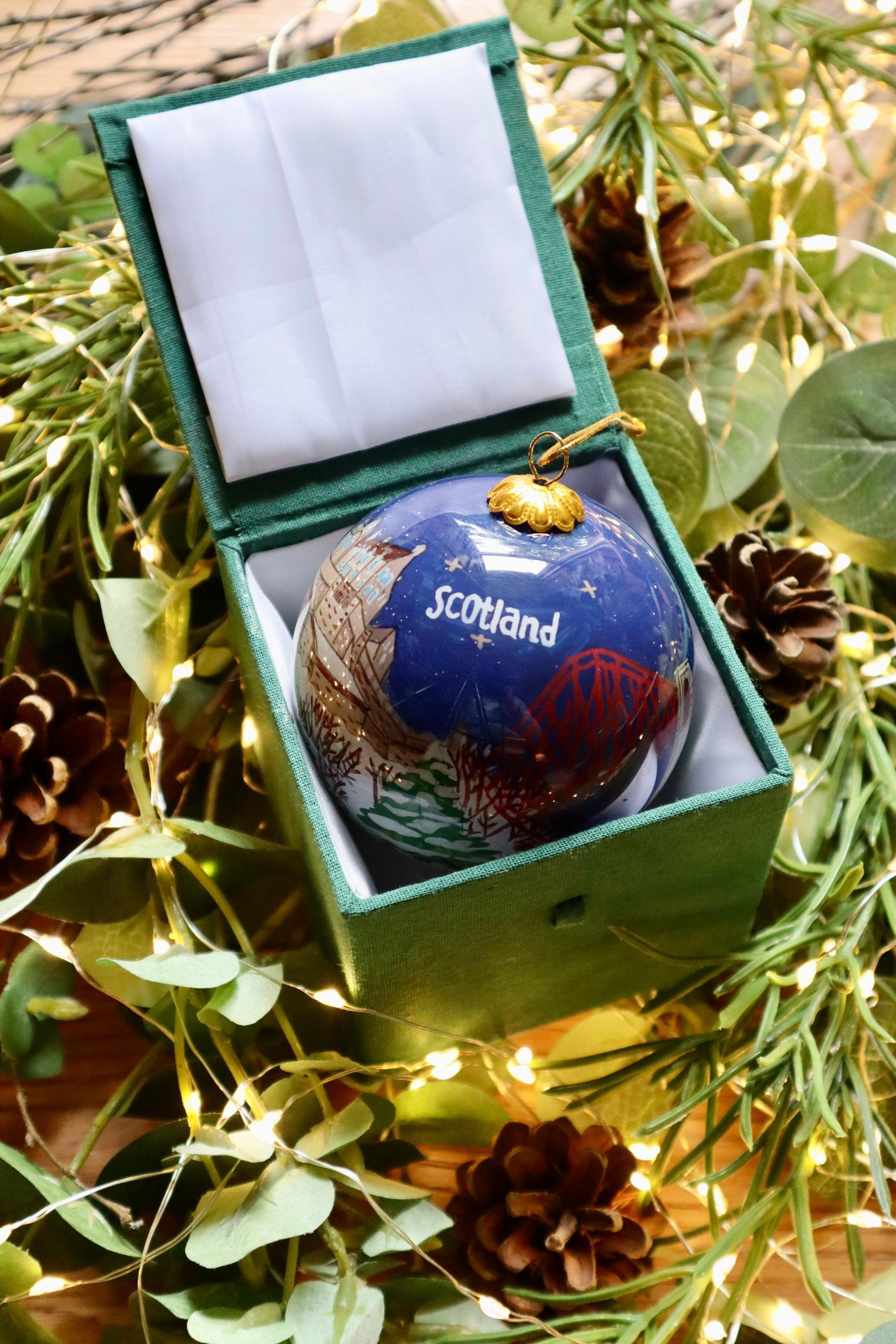 A glossy blue glass christmas bauble featuring a detailed map of Scotland with red and gold accents in a velvet box
