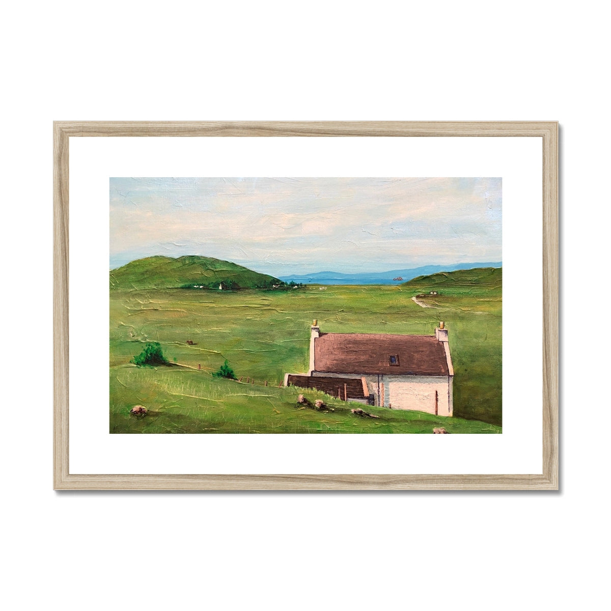 Framed landscape print of a white house with brown roof in a green field near hills and water