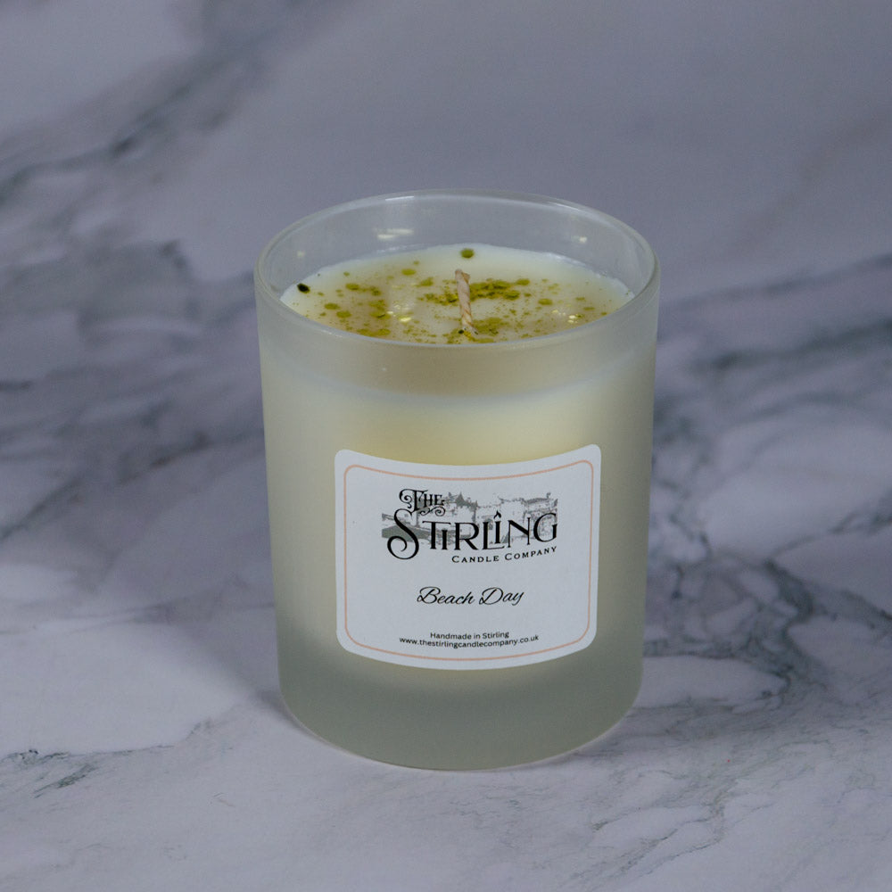 A frosted glass medium candle jar with creamy white wax and green flecks labeled beach day by the string candle company
