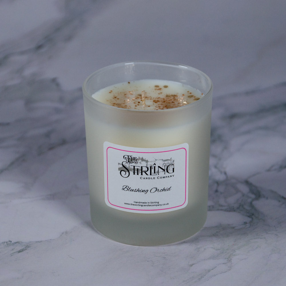 A frosted glass medium candle jar with creamy white wax and golden flecks labeled blushing orchid by Stirling Candle Company