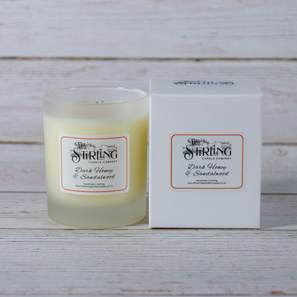 A creamy yellow scented candle in a frosted glass jar labeled dark honey and sandalwood by the stirling candle company