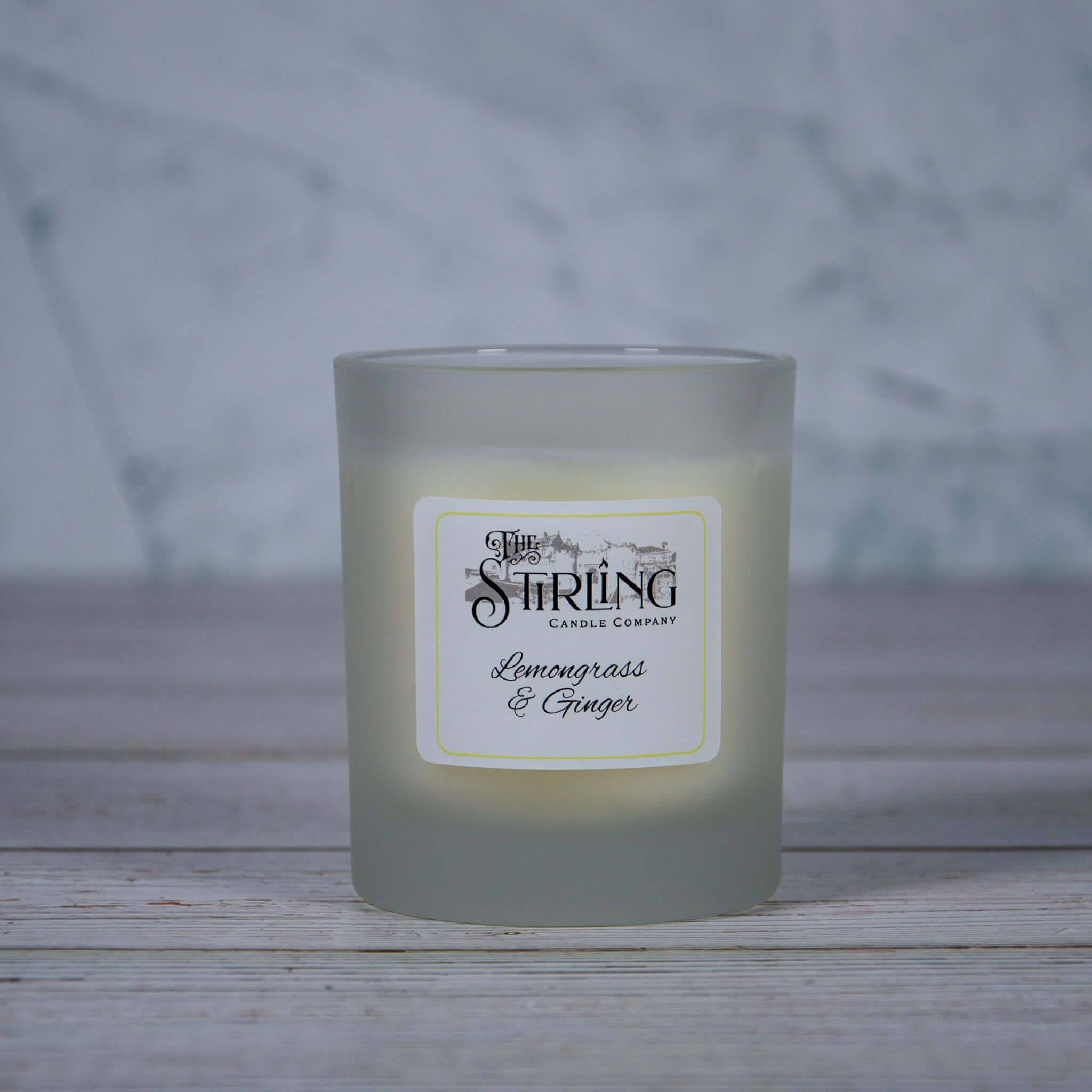 A frosted glass candle jar with a white label featuring lemongrass and ginger by the string candle company medium candle