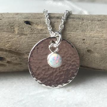 Silver circular pendant with a hammered texture, featuring a small opal stone, hanging from a twisted chain on a natural wood