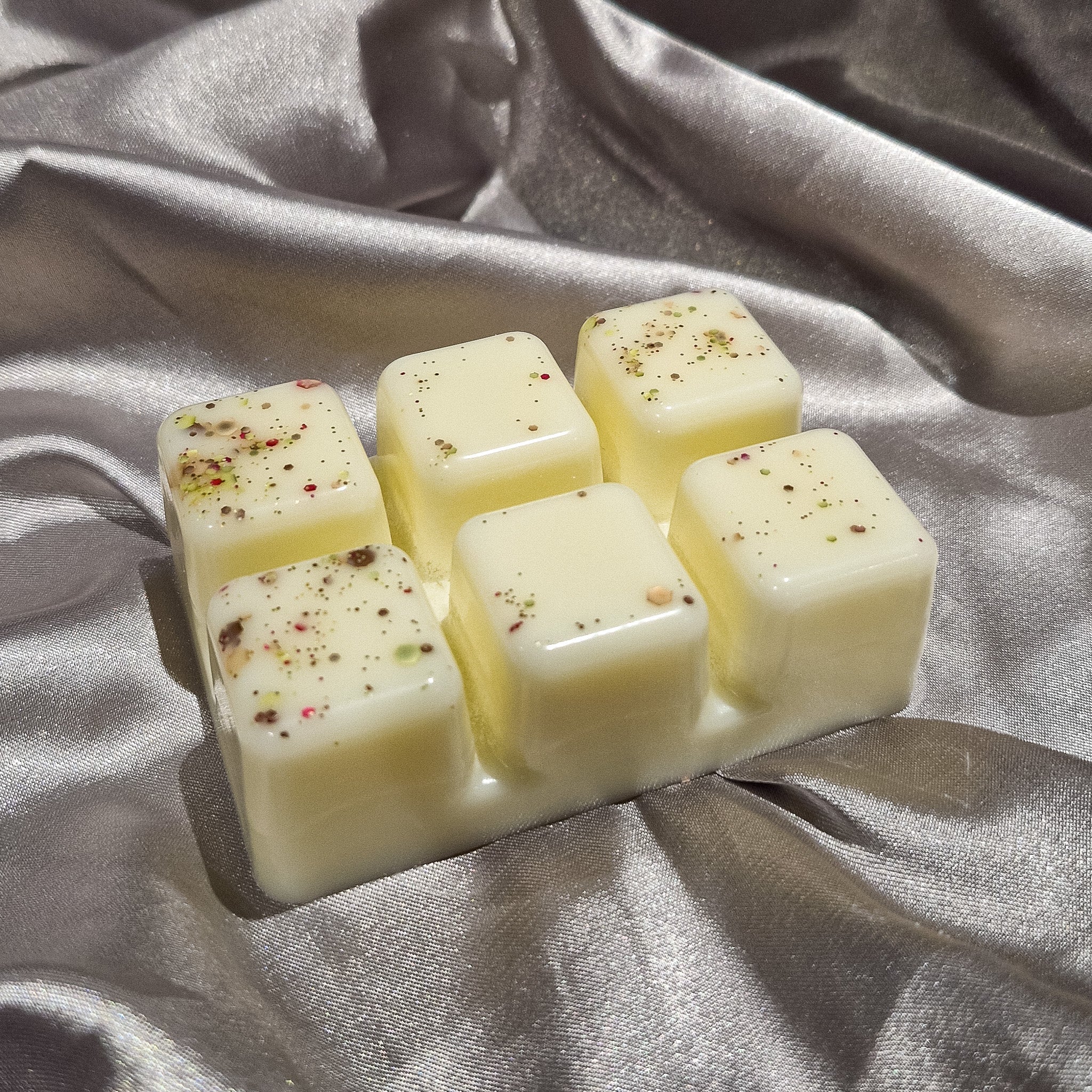 Six pale yellow dark honey and sandalwood wax melts with green red and brown flecks in a molded clamshell tray
