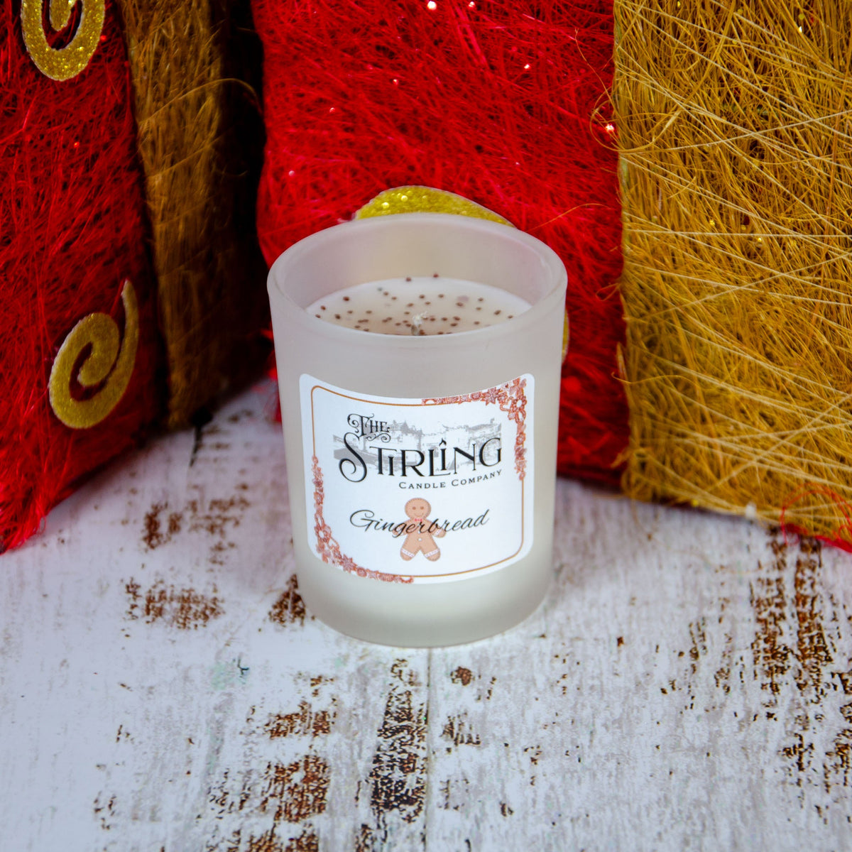 Frosted glass candle with a gingerbread scent label, placed on a rustic wooden surface near decorative red and gold items.
