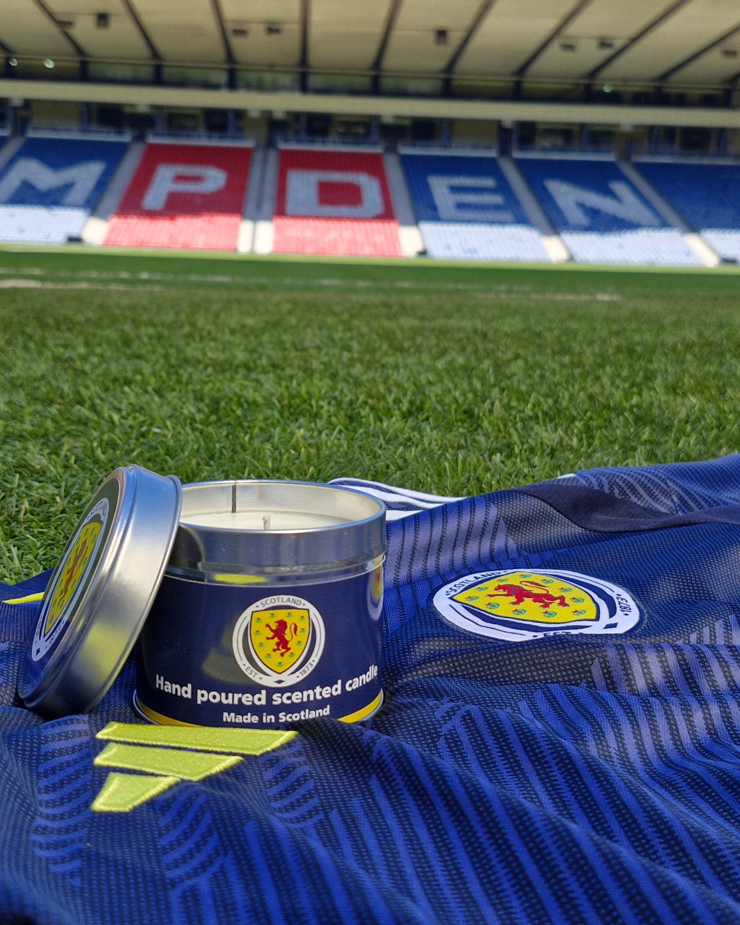 A hand-poured scented candle in a metallic tin with a blue label featuring the scottish emblem on a royal blue football jersey
