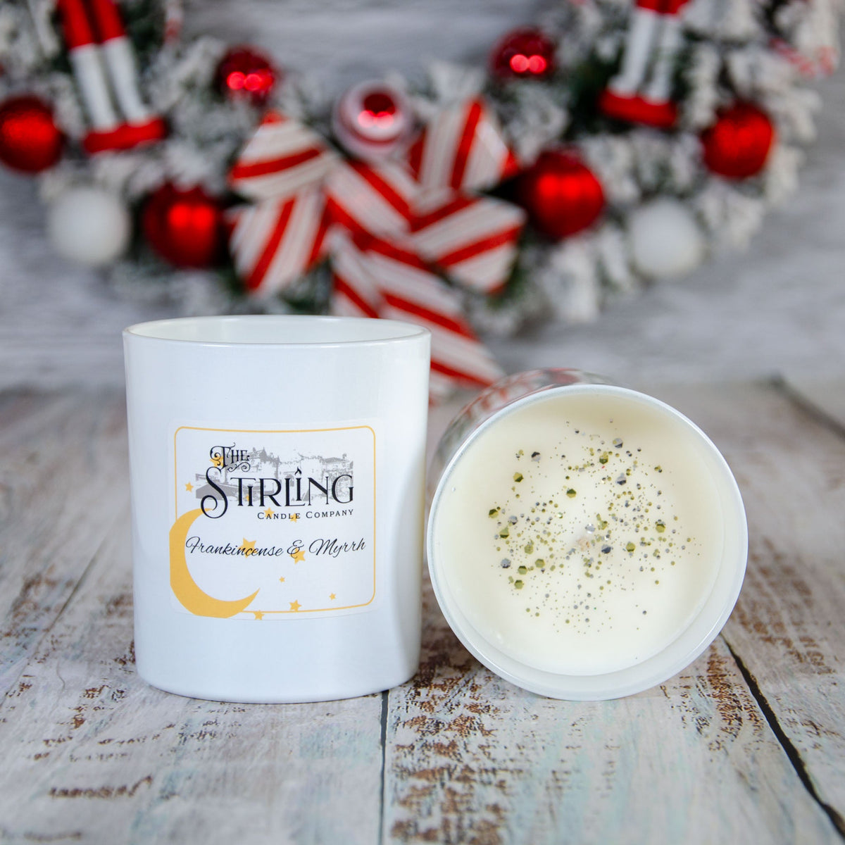 White candle with a logo, placed next to a festive wreath with red and white decorations.