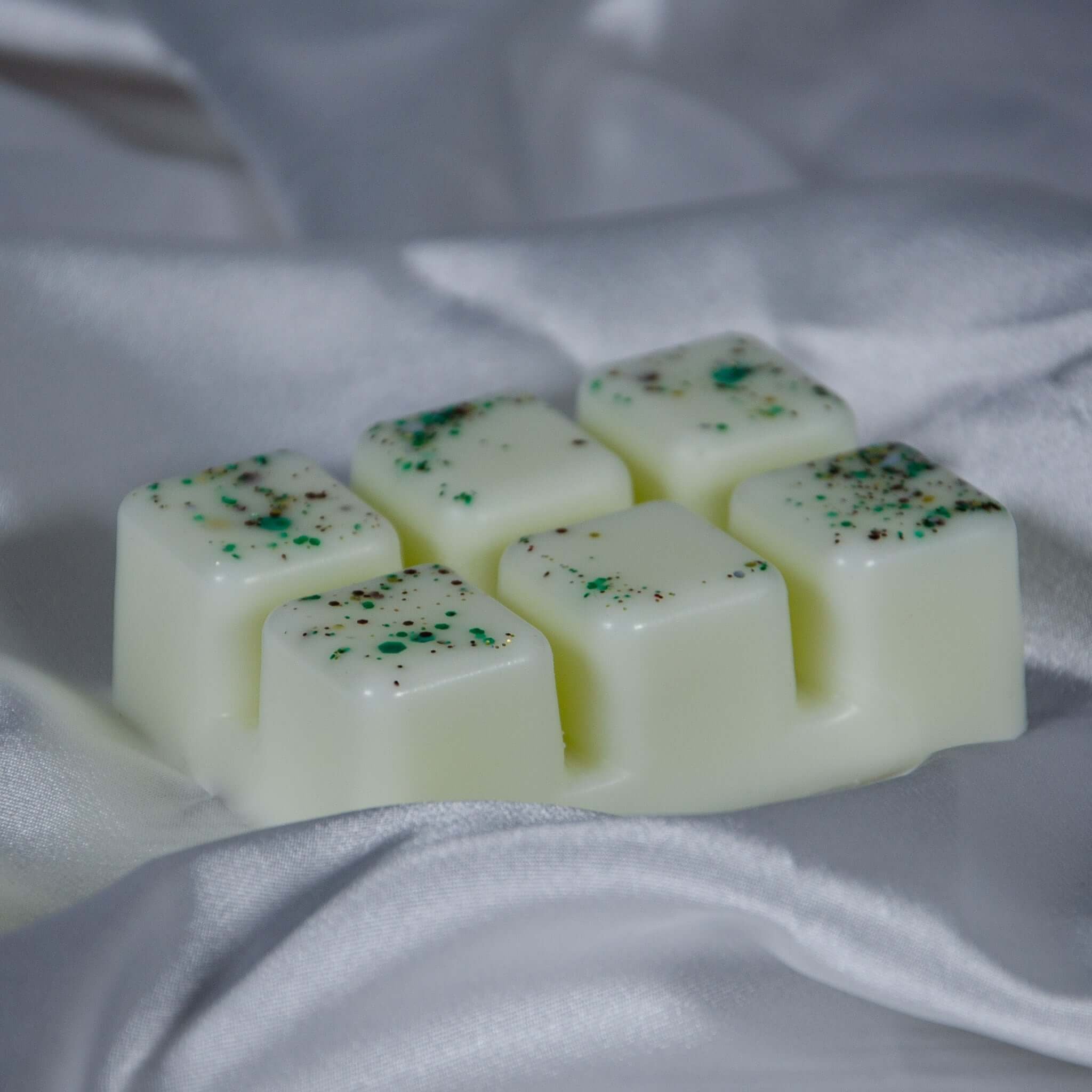 Six pale green glitter-speckled wax melts in Lemongrass & Ginger clamshell on white fabric