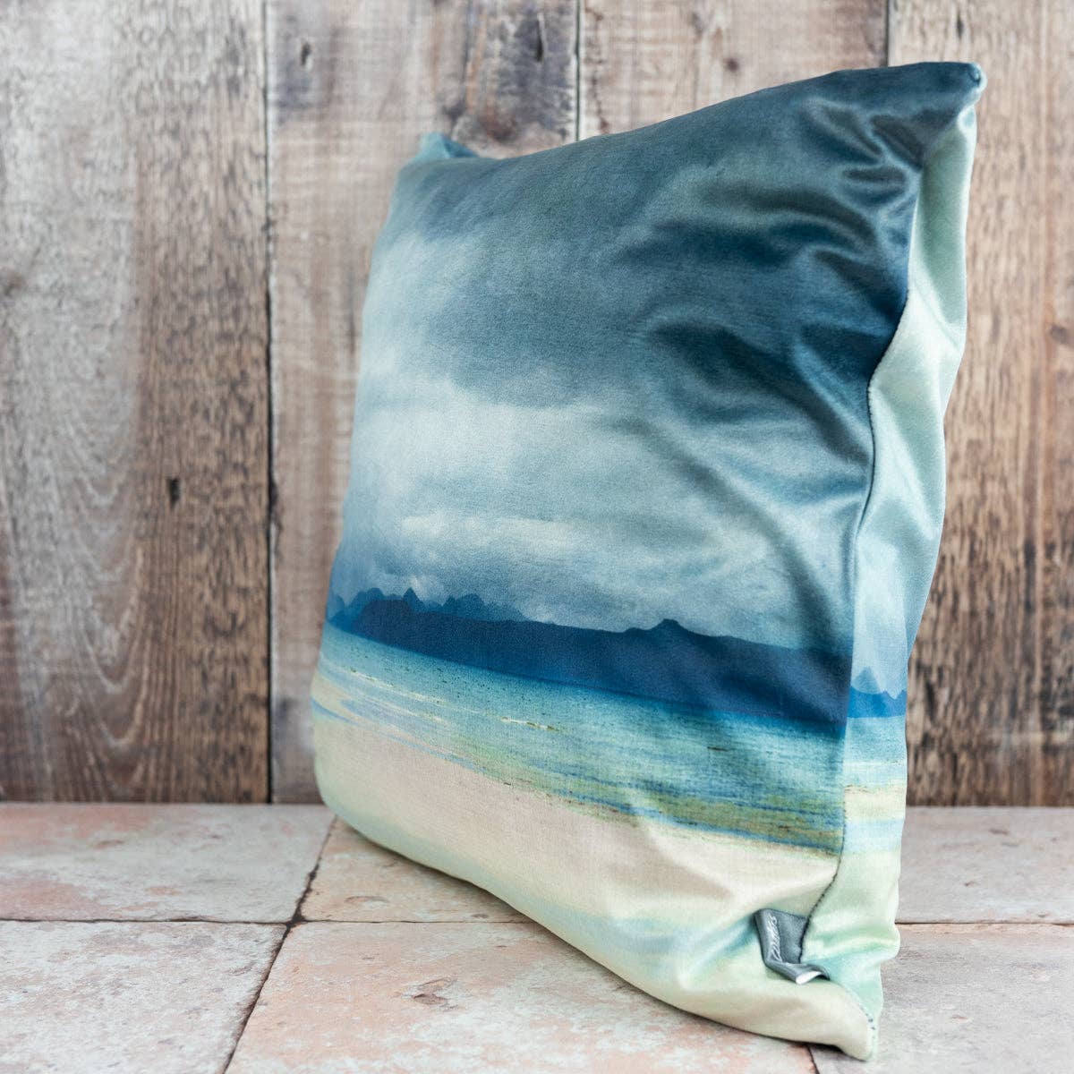 A square satin cushion from applecross velvet featuring an abstract coastal scene in blue and teal shades