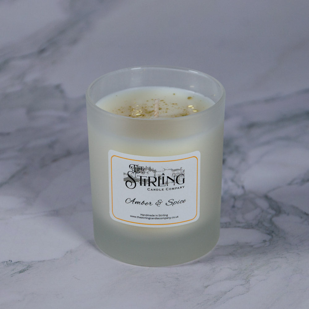 A frosted glass medium candle jar with white wax and golden flecks labeled amber and spice by the Stirling Candle Company