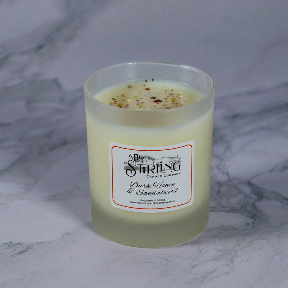 A frosted glass medium candle jar with creamy yellow wax and speckled top labeled dark honey and sandalwood by stirling candle company