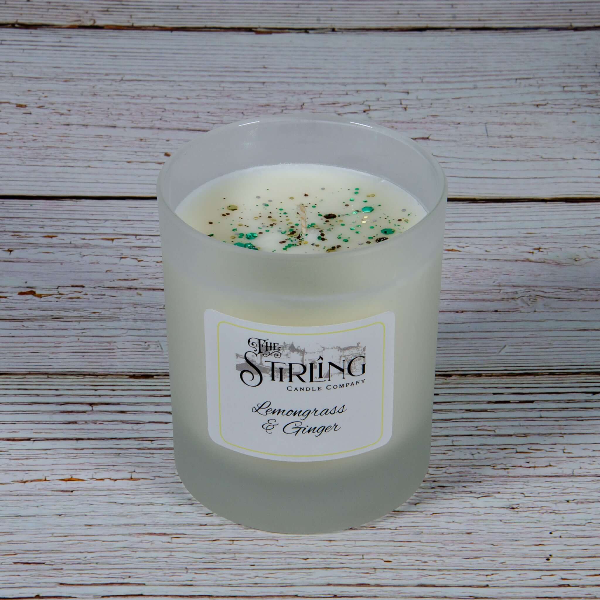 A frosted glass medium candle jar with white wax speckled green and black labeled lemongrass and ginger by the Stirring Candle Company